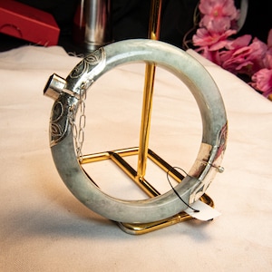 May include: A light green jade bangle bracelet with silver accents. The bracelet features ornate silver detailing on the clasp and end pieces, connected by a delicate chain. The jade has a subtle, natural color variation.