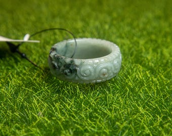 May include: A light green jade ring with carved floral patterns. The ring has a band of swirling designs and a darker green area. The ring is set against a backdrop of green grass.
