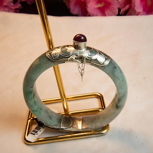 Puede incluir: Un brazalete de jade con detalles plateados y una pequeña gema roja. El brazalete se exhibe en un soporte dorado. El jade tiene un aspecto verde moteado.