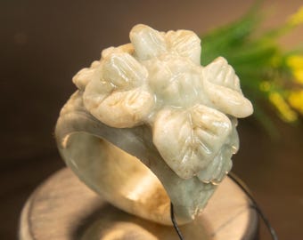 以下が含まれることがあります： 淡い緑色の翡翠の指輪、花の模様。 指輪は、詳細な花びらと葉を持つ彫刻された花が特徴です。 指輪は銀色の表面に置かれ、ジュエリーの複雑な職人技を紹介しています。