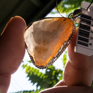 May include: A pendant featuring a translucent, irregularly shaped stone with orange and white hues, set in a silver-colored metal frame. The stone has a unique, natural pattern. The pendant is suspended from a thin silver chain.