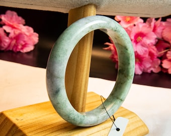 Op de afbeelding: Een jade armband met een lichtgroen en wit gemarmerd patroon. De armband wordt gepresenteerd op een houten standaard met een grijze stoffen bovenkant. Roze bloemen zijn zichtbaar op de achtergrond.