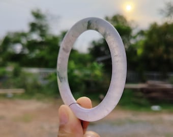 May include: A translucent, lavender-colored jade bangle bracelet. The smooth, polished surface of the bracelet reflects the sunlight. The jade has subtle green inclusions. The bracelet is held up against a blurred background of trees and a setting sun.