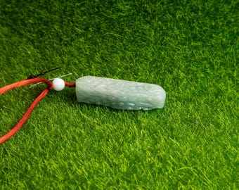 Puede incluir: Un colgante de jade verde claro con una superficie texturizada, de forma rectangular, suspendido por un cordón rojo. Una cuenta blanca conecta el colgante al cordón. El colgante está sobre un fondo verde.