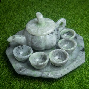 Könnte beinhalten: Ein Jade-Teeservice mit einer Teekanne mit Deckel, einem Tablett und sechs kleinen Tassen. Das Set ist hellgrün mit dunkleren grauen und schwarzen Adern. Die Teekanne hat einen gebogenen Griff und Ausguss. Das Set steht auf einer grünen Oberfläche.