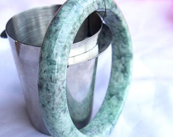 Op de afbeelding: Een jade armband met een gevlekt groen en wit patroon. De armband is cirkelvormig en bevindt zich in de buurt van een kleine, zilveren metalen emmer. De achtergrond is zacht wit en roze.