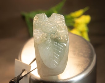 May include: A carved jade pendant in shades of pale green and white. The pendant features a floral design with detailed leaf patterns. The jade is translucent, and the carving is intricate. The pendant is displayed on a silver-colored surface.