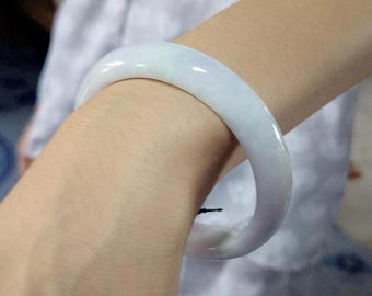 Op de afbeelding: Een lichtpaarse jade armband. De gladde, gepolijste armband heeft een lichtpaarse kleur en wordt om de pols gedragen. De armband heeft een solide, cirkelvormige vorm en lijkt gemaakt van een doorschijnende steen.