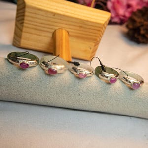 May include: Five oval-shaped pendants with silver-colored metal bands and a small, round, pink gemstone. Two pendants are green, and three are clear. Each pendant has a black cord attached.