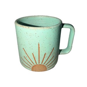 May include: A ceramic mug with a light teal glaze and a speckled finish. The mug features a brown sunburst design on the front. It has a rounded handle and a cylindrical shape.