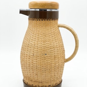 Op de afbeelding: Een vintage Corning Designs thermoskan. De kan heeft een geweven, lichtbruine buitenkant met een donkerbruine basis, rand en tuit. Het heeft een houten deksel en een gebogen handvat. De kan is ongeveer 30 cm hoog.