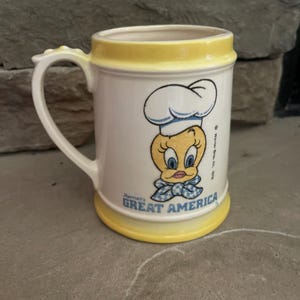 May include: A white ceramic mug with a yellow rim and base, featuring a cartoon image of Tweety Bird wearing a chef's hat and a blue and white checkered bow. The mug has a curved handle and the text "Mamall's GREAT AMERICA".