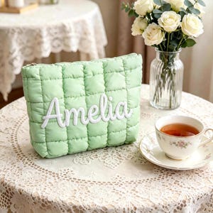 Puede incluir: Un neceser acolchado verde claro con el nombre "Amelia" bordado en blanco. El neceser es rectangular y tiene cierre de cremallera. Una taza de té y un jarrón con rosas blancas están sobre la mesa.