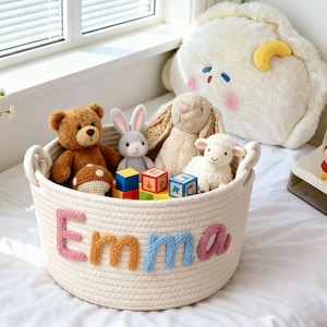 May include: A cream-colored rope basket filled with stuffed animals and wooden blocks. The name "Emma" is spelled out in colorful yarn letters on the basket. A teddy bear, bunny, and lamb are visible.