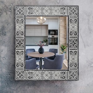 May include: Rectangular mirror with a decorative black and white patterned frame. The frame features geometric and scroll designs. The mirror reflects a dining area with a round table, chairs, and a vase of flowers.