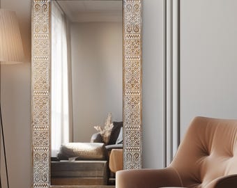 May include: A rectangular full-length mirror with a decorative, patterned frame. The frame is a light brown color with white geometric designs. The mirror reflects a room with a chair and a lamp. The mirror is a decorative home decor item.
