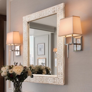 May include: A rectangular mirror with a decorative, patterned frame and two matching sconces. The sconces have rectangular shades and are mounted on the wall. A vase of flowers sits below the mirror.
