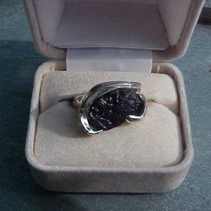 May include: A silver ring with a black stone set in a unique design. The ring is displayed in a white ring box.