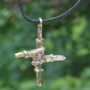 Puede incluir: Un colgante de color dorado en forma de cruz con un diseño de nudo celta. El colgante está colgado de un cordón negro.