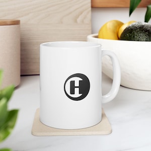 Puede incluir: Taza de cerámica blanca con una gran letra "H" negra dentro de un círculo negro. La taza tiene un asa curva y se apoya en un posavasos beige. El fondo incluye una superficie de madera y un cuenco blanco con fruta.
