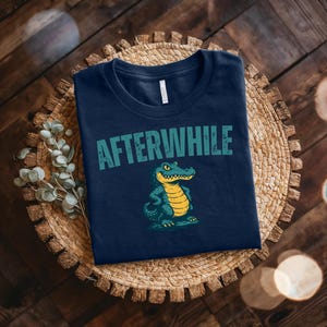 May include: Navy blue t-shirt with the text "AFTERWHILE" in teal above a cartoon alligator illustration. The alligator is teal and yellow. The shirt is folded on a woven, circular mat.