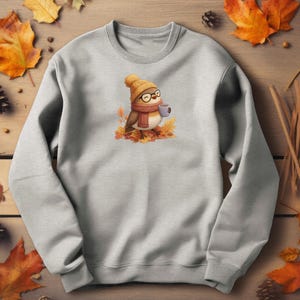 Op de afbeelding: Een lichtgrijs sweatshirt met een cartoonvogel met een bril, een hoed en een sjaal, die een mok vasthoudt. De vogel is omringd door herfstbladeren. Het ontwerp is gecentreerd op de borst van de sweatshirt.