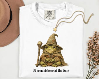 Op de afbeelding: Wit T-shirt met een illustratie van een kikker als tovenaar, met een puntmuts en staf. De tekst "It seemed wise at the time" staat onder de kikker. Een gouden sterrenketting en een bruine hoed zijn ook zichtbaar.