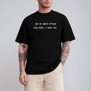 May include: Black t-shirt with the text "ASK ME ABOUT PYTHON CODE BUGS, I DARE YOU" in white. The shirt is a crew neck and has short sleeves. The person is wearing khaki pants.