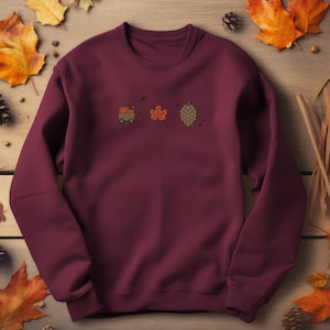 Puede incluir: Sudadera burdeos de cuello redondo con diseños bordados con temática otoñal. El diseño incluye un carro lleno de calabazas, una hoja y una piña. La sudadera está rodeada de hojas de otoño y piñas.