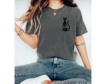 Pode incluir: T-shirt cinza escuro com uma silhueta de gato preto e um design de paisagem urbana. O gato tem olhos verdes e está sentado em posição vertical. A camisa tem um ajuste folgado e mangas curtas.