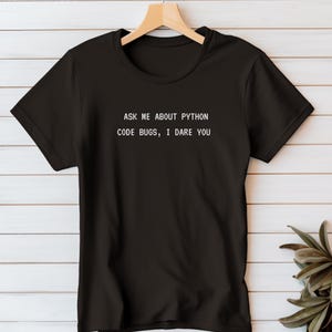 May include: Black t-shirt with the text "ASK ME ABOUT PYTHON CODE BUGS, I DARE YOU" in white. The shirt is hanging on a wooden hanger against a white wood-paneled wall. A green plant is visible in the lower right corner.