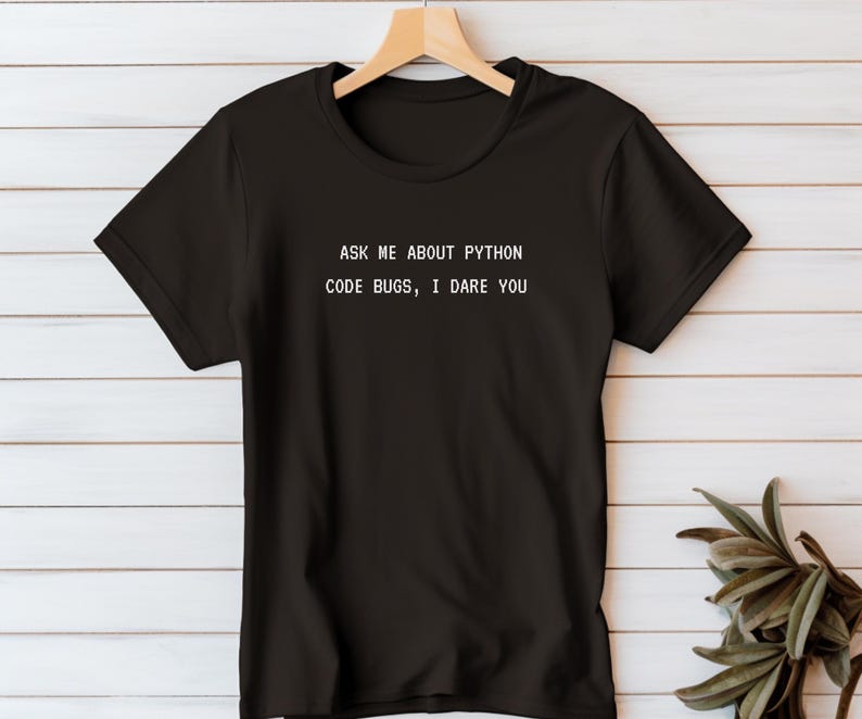 May include: Black t-shirt with the text "ASK ME ABOUT PYTHON CODE BUGS, I DARE YOU" in white. The shirt is hanging on a wooden hanger against a white wood-paneled wall. A green plant is visible in the lower right corner.