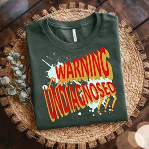Puede incluir: Camiseta verde oscuro con el texto "WARNING I'M UNDIAGNOSED" en letras rojas y amarillas. La camiseta está doblada y colocada sobre un salvamanteles circular tejido. El texto tiene un efecto desgastado y salpicado.