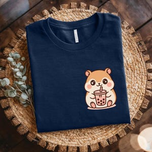 May include: Navy blue t-shirt featuring a cartoon hamster holding a bubble tea. The hamster is light brown with pink cheeks and a white belly. The bubble tea is pink with a brown straw. The t-shirt is displayed on a woven placemat.