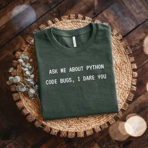 May include: Dark green t-shirt with the text "ASK ME ABOUT PYTHON CODE BUGS, I DARE YOU" in white. The shirt is laid flat on a woven, circular placemat. The background is a dark wooden surface.