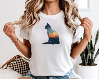 Pode incluir: T-shirt branca com silhueta de gato. O gato é azul escuro e a paisagem urbana no interior tem tons de amarelo, laranja e azul. Uma peça casual para o dia a dia.