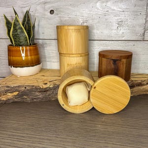May include: A collection of small, round bamboo containers, some stacked, with a natural wood finish. One container is open, revealing a small, square, off-white fabric pouch inside. A small, potted plant with green and yellow leaves is in the background.