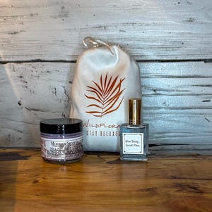 May include: A collection of self-care products, including a jar of Modern Skyn Alchemy Brazilian Clay, a bottle of Blue Taney Facial Mist, and a drawstring bag with the Wildflora logo. The products are arranged on a wooden surface.