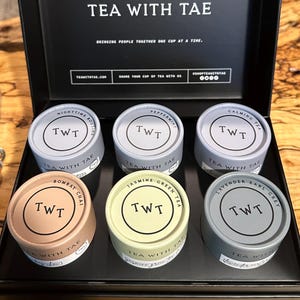 May include: A black box containing six tea tins. Flavours include Nighttime Routine, Peppermint, Calming Tea, Bombay Chai, Jasmine Green Tea, and Lavender Earl Grey. The brand name "TEA WITH TAE" is printed on the box and tins.