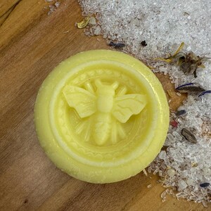 Peut inclure: Un galet de bain jaune pâle avec un motif d'abeille détaillé au centre. Le galet est entouré de cristaux blancs et de plantes séchées sur une surface en bois. L'abeille est le point central.