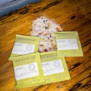 May include: Four green tea sachets with the text "TEA WITH TAE" and "genmaicha" are arranged on a wooden surface, alongside a pile of loose tea leaves and flower petals. The sachets list ingredients and caffeine levels.