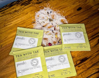 May include: Four light green tea sachets labeled Tea With Tae Genmaicha with green tea leaves and rice ingredients, arranged around a central pile of loose tea blend containing white salt crystals, dried flower petals, and green tea leaves.