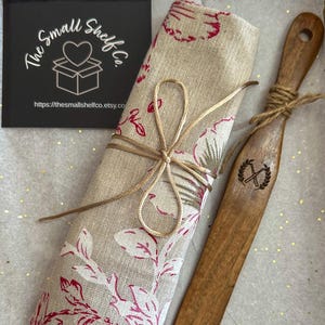 May include: A rolled linen kitchen towel with a red floral pattern is tied with twine, next to a wooden spatula. A black card with the text "The Small Shelf Co." and a heart logo is also visible.