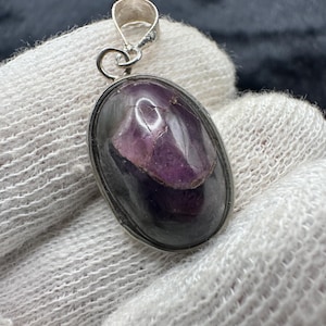 May include: An oval-shaped pendant featuring a polished, dark purple and black stone set in a silver-toned metal bezel. The stone has a smooth, reflective surface with visible natural inclusions and color variations. A silver-toned metal bail is attached.