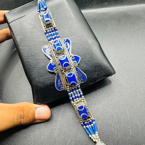 May include: A silver and blue bracelet with a central butterfly-shaped design. The bracelet features square blue stones and blue beaded strands. The design is set against a black background.