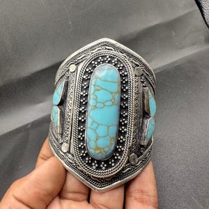 May include: A wide silver cuff bracelet with turquoise stones. The bracelet has an ornate design with a large oval turquoise stone in the centre and smaller stones on the sides. The metal has a detailed, textured pattern.