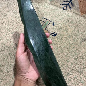 May include: A long, smooth, dark forest green stone object with a polished, reflective surface. The item features a slightly curved, elongated shape with a rounded top and a wider, flat base, showcasing natural mottled patterns within the stone.