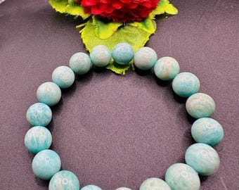 Könnte beinhalten: Ein türkisfarbenes Perlenarmband mit Marmormuster. Das Armband besteht aus runden Perlen und wird auf einer dunklen Oberfläche präsentiert. Eine rote Blume mit grünen Blättern befindet sich im Hintergrund.