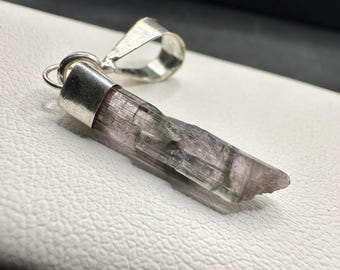 May include: A natural gemstone pendant with a silver bail. The gemstone is a light purple colour with a crystalline structure. The pendant is set in a silver cap. The pendant is displayed on a white surface.
