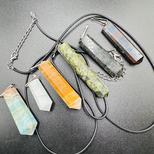 May include: Assortment of gemstone pendants on black cords. The pendants are various colors, including blue, orange, green, and brown. Each pendant is a rectangular shape with a pointed end. Some pendants have silver chain accents.